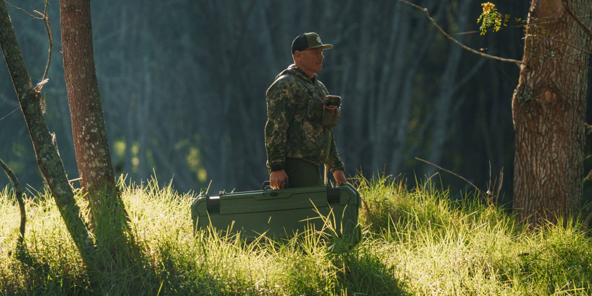 A man in camo walking through the woods, carrying his Ranger Green Honcho 80 D-co case that he uses for his bow.