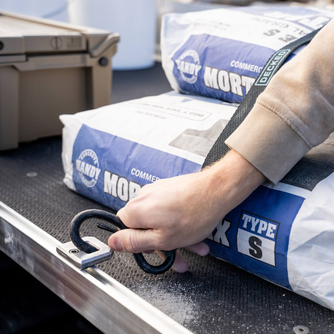 A man attaching a cam strap to a D-Ring Tie Down to hold a stack of mortar mix bags to the CargoGlide deck.