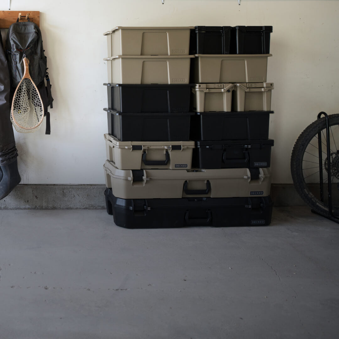 A mix of colors and sizes of D-Co Cases stacked in a garage with fishing waders and a bike sitting nearby.