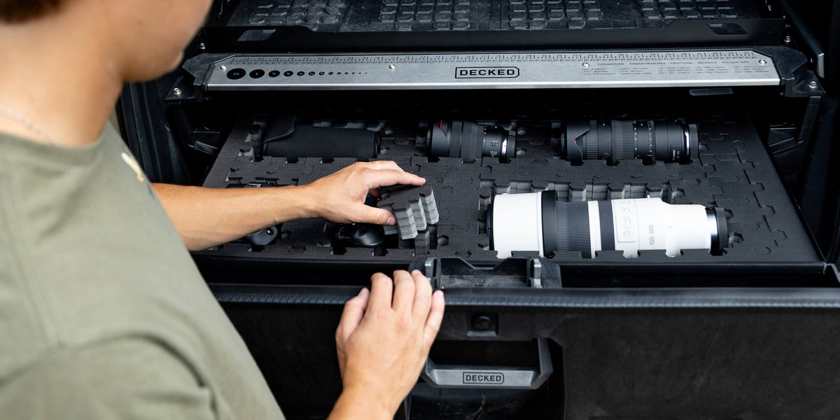 Close-up of a person removing interlocking foam pieces from a DECKED Drawer System to create a custom cutout, revealing camera lenses and gear nested in the puzzle foam, with a DECKED ruler on the drawer's front rail."