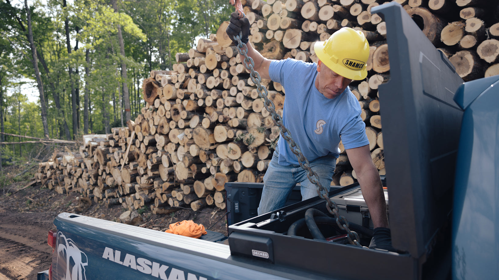 Timber worker storing a heavy logging chain in a DECKED Tool Box beside stacked logs.
