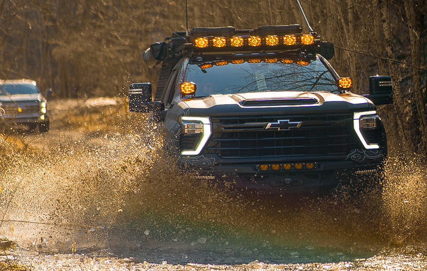 Chevrolet Silverado overland truck driving through a shallow water crossing with roof-mounted lights and off-road accessories.