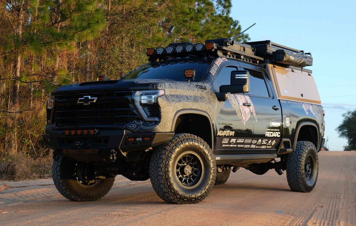 Lifted Chevrolet Silverado overland truck with a truck canopy, roof rack, off-road tires, and auxiliary lighting parked on a dirt road.