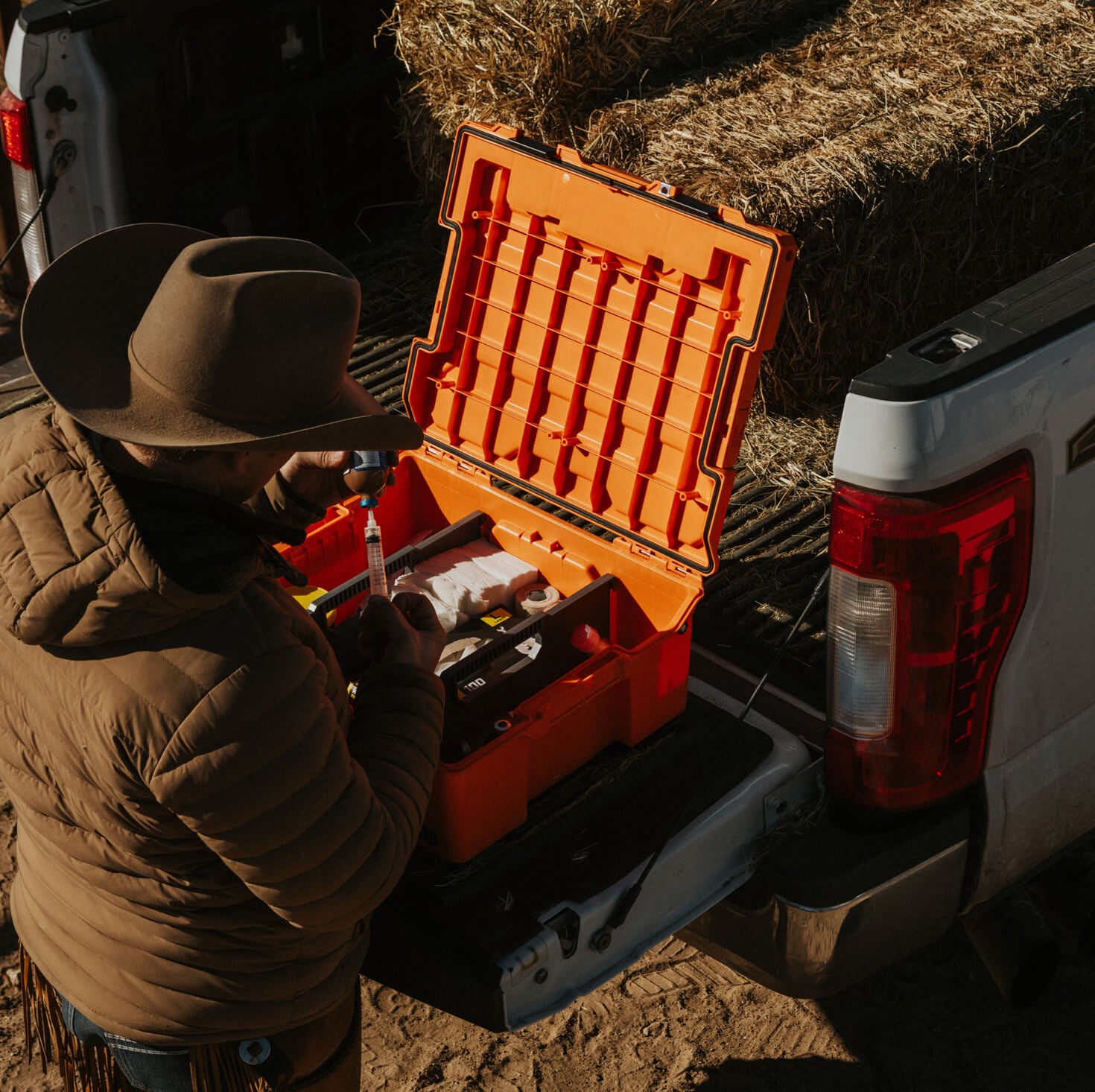 Person in a cowboy hat using tools from an orange Halfrack 32 case in a truck bed with hay.