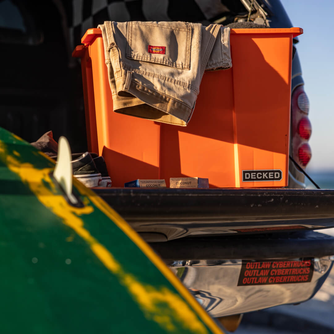 Orange Payloader 64 sitting on a tailgate with a folded pair of work pants hanging out and a surfboard sitting nearby.