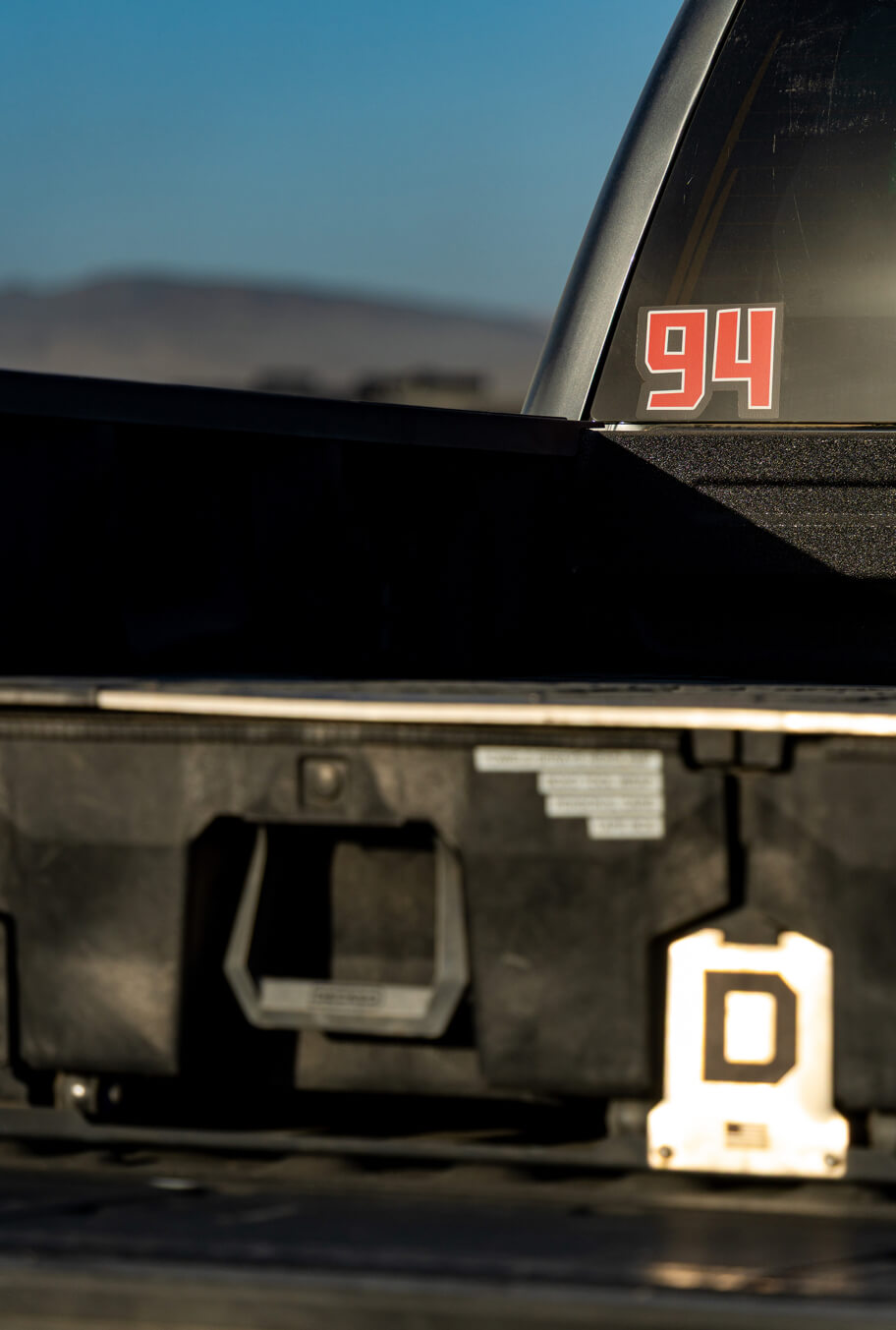 Trunk of a vehicle with a '94' decal on the rear window, set against a blurred natural background.