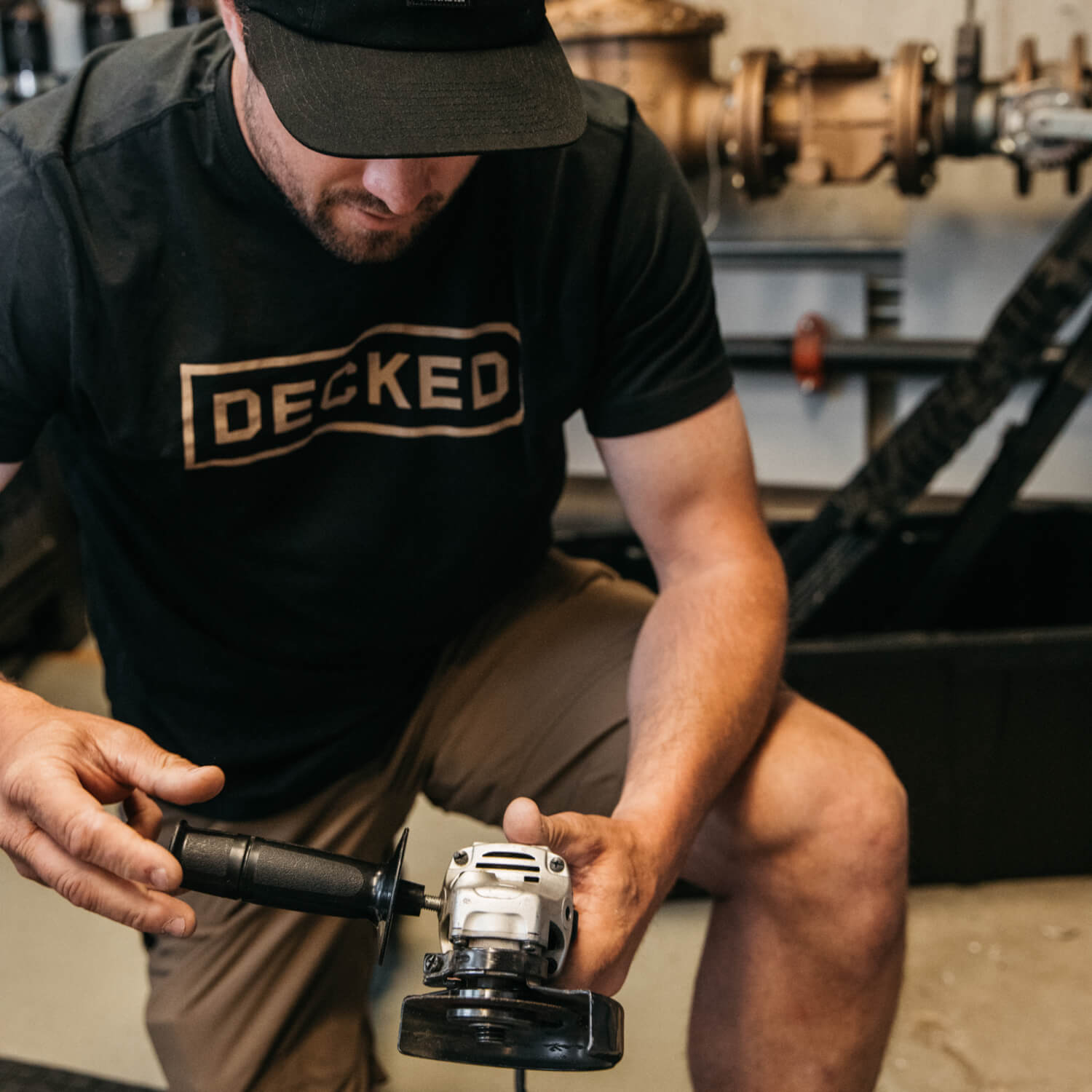 [T0001-DECK-BLK, T0002-DECK-BLK, T0003-DECK-BLK, T0004-DECK-BLK, T0005-DECK-BLK, T0006-DECK-BLK] Man in a black 'DECKED' shirt using a power tool in a workshop setting.
