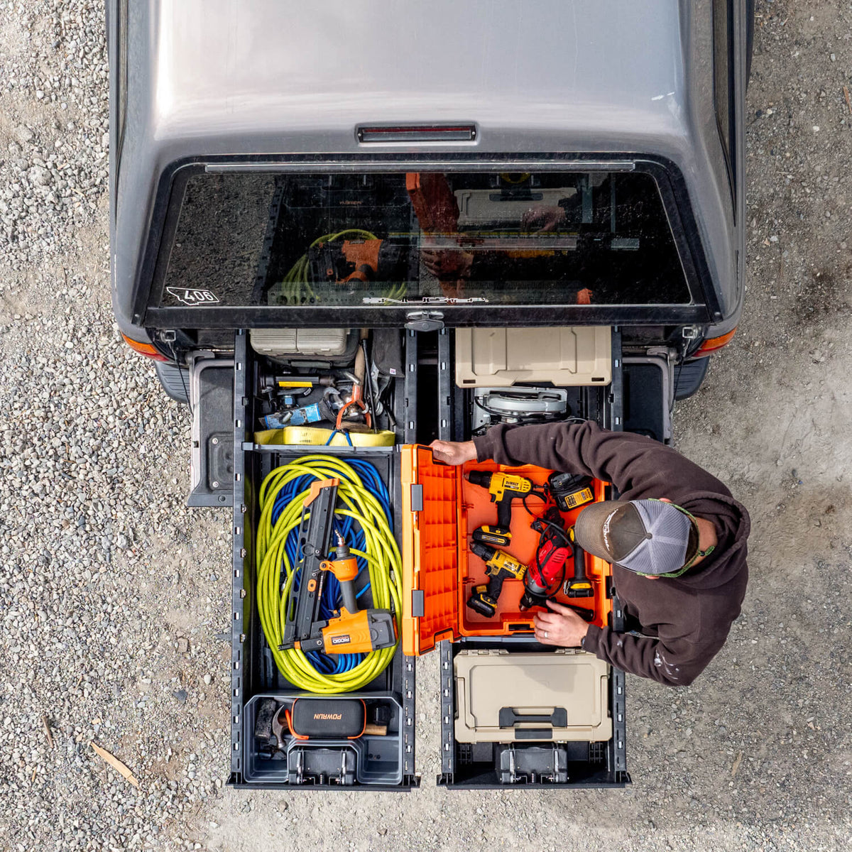 Top-down view of a person organizing tools in the open tailgate with a Drawer System extended out of a gray pickup truck. The Drawer System contains organized storage compartments with orange tool bins, yellow extension cords, power tools, and tan storage cases on a gravel surface.