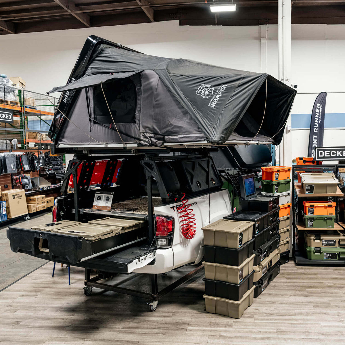 A black rooftop tent mounted on the roof of a white pickup truck, shown in a warehouse or showroom with storage bins and shelving in the background. The tent has a visible logo on the front panel.