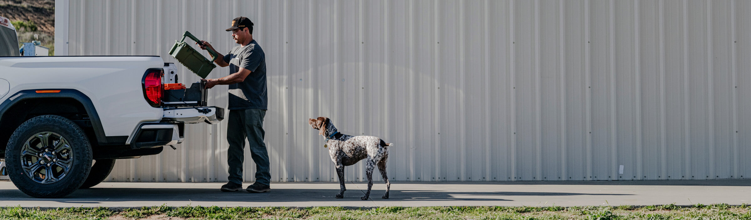 Man loading a protective case into a truck bed's DECKED Drawer System with a dog standing nearby, against a gray building.