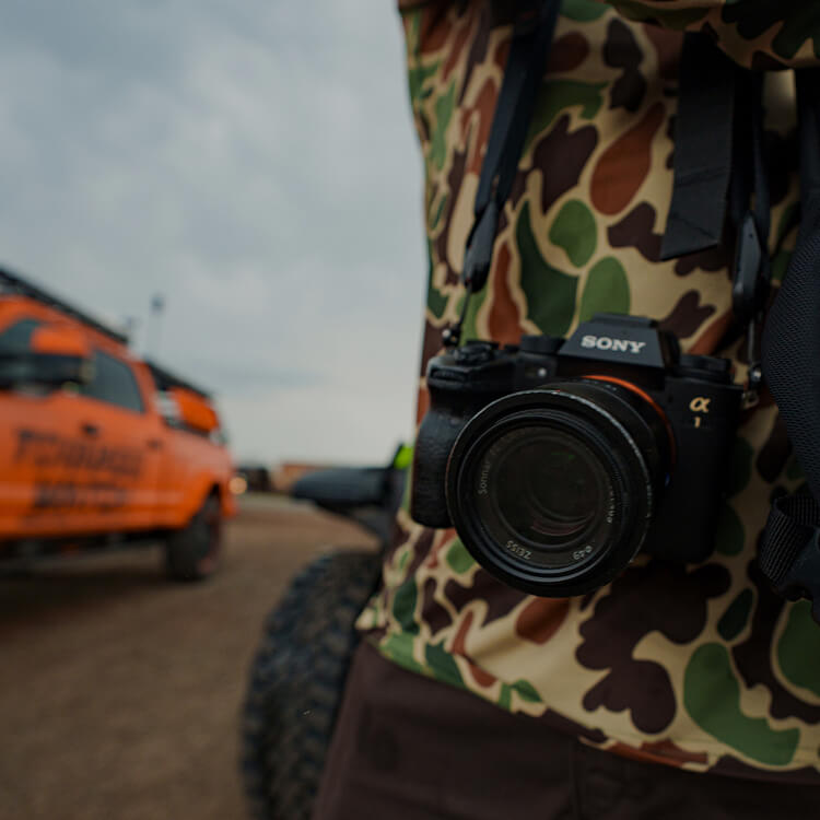 Person wearing a camouflage shirt with a Sony camera on a dirt road with vehicles in the background