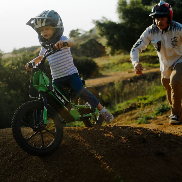 A little girl riding a green balance bike on a dirt single track with her dad chasing after her.