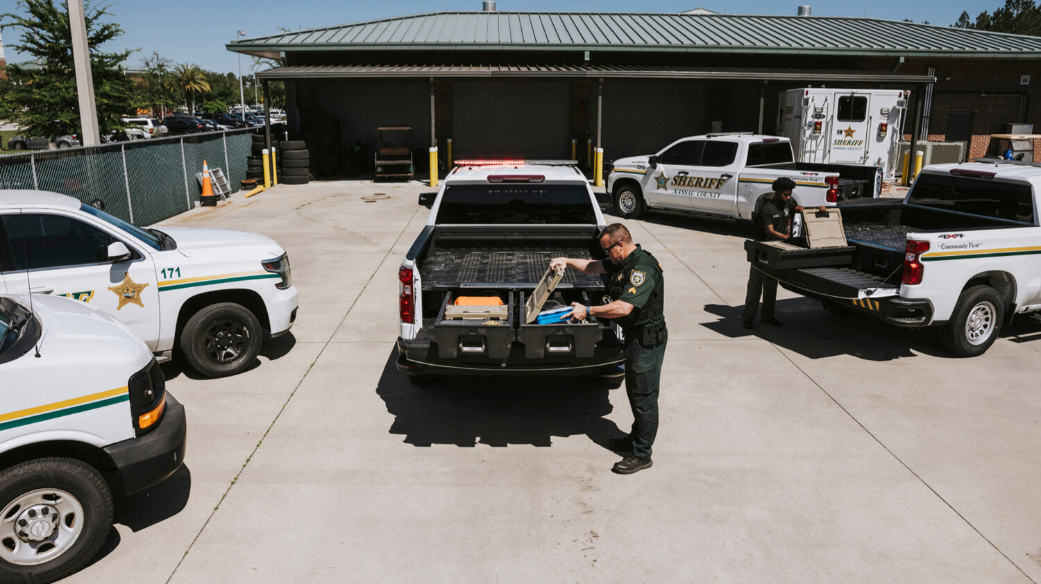 Law enforcement officers retrieving gear from their DECKED Drawer System in a truck in a parking lot with other vehicles and a building in the background.