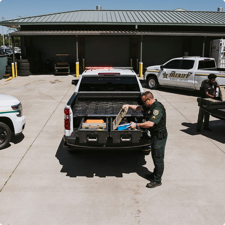 Law enforcement officer loading equipment into a truck in a parking lot with other vehicles and a building in the background.