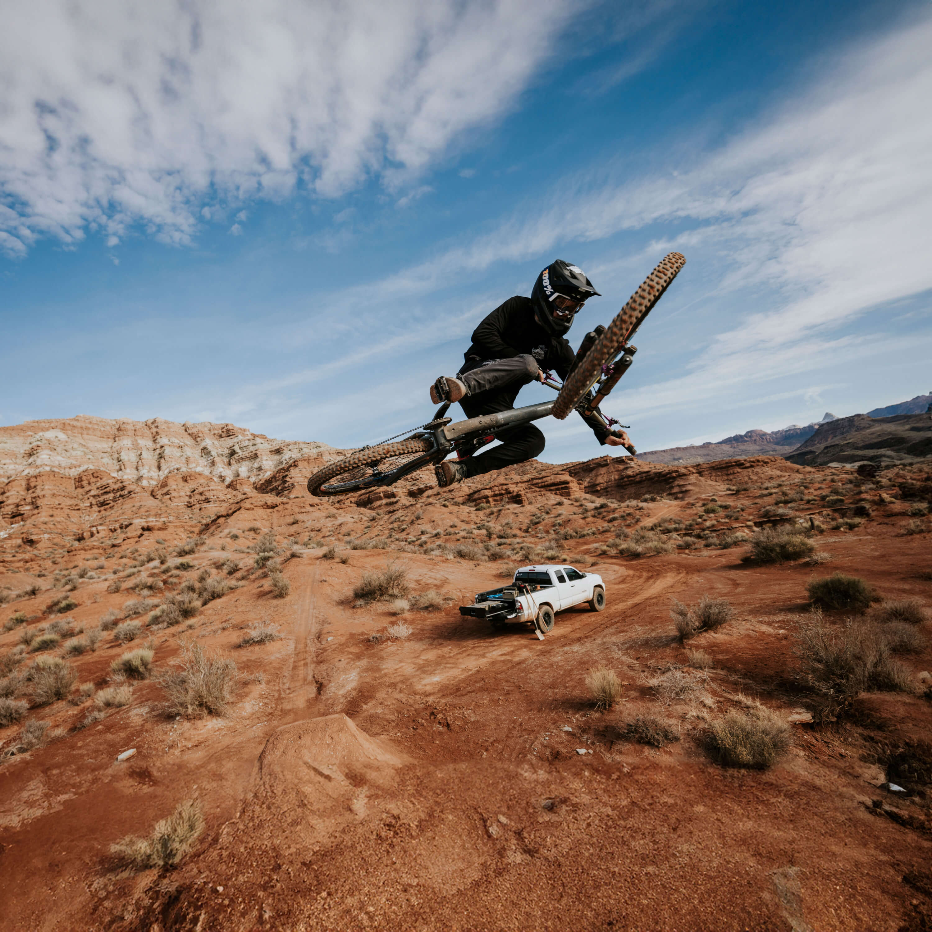 A biker in the red rocks mid air. In the distance, you can see his truck with his DECKED Drawer System open to reveal his organized D-Co cases.