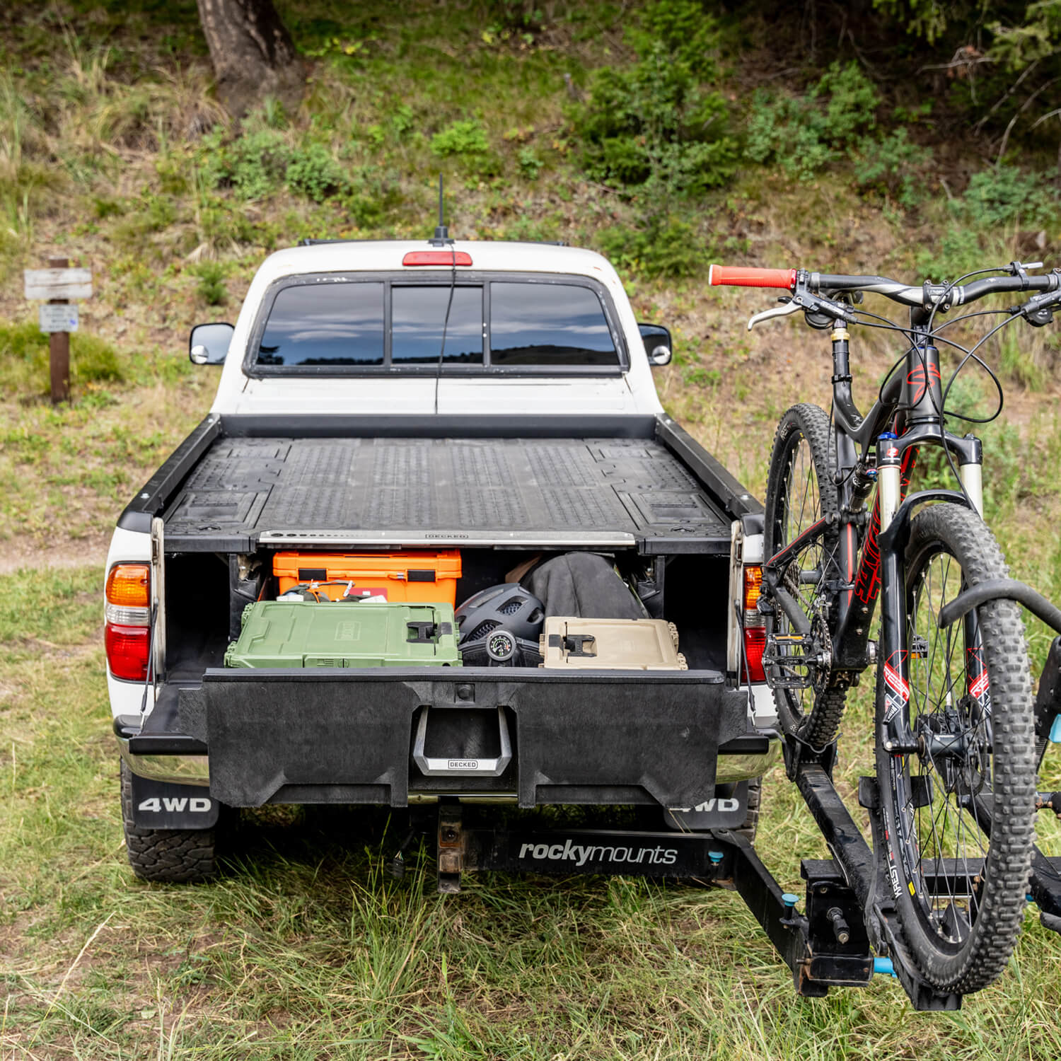 A white Gen 1 Tacoma with a bike trailer attached, parked on a dirt road with a Drawer System open to reveal D-co cases in the truck bed.