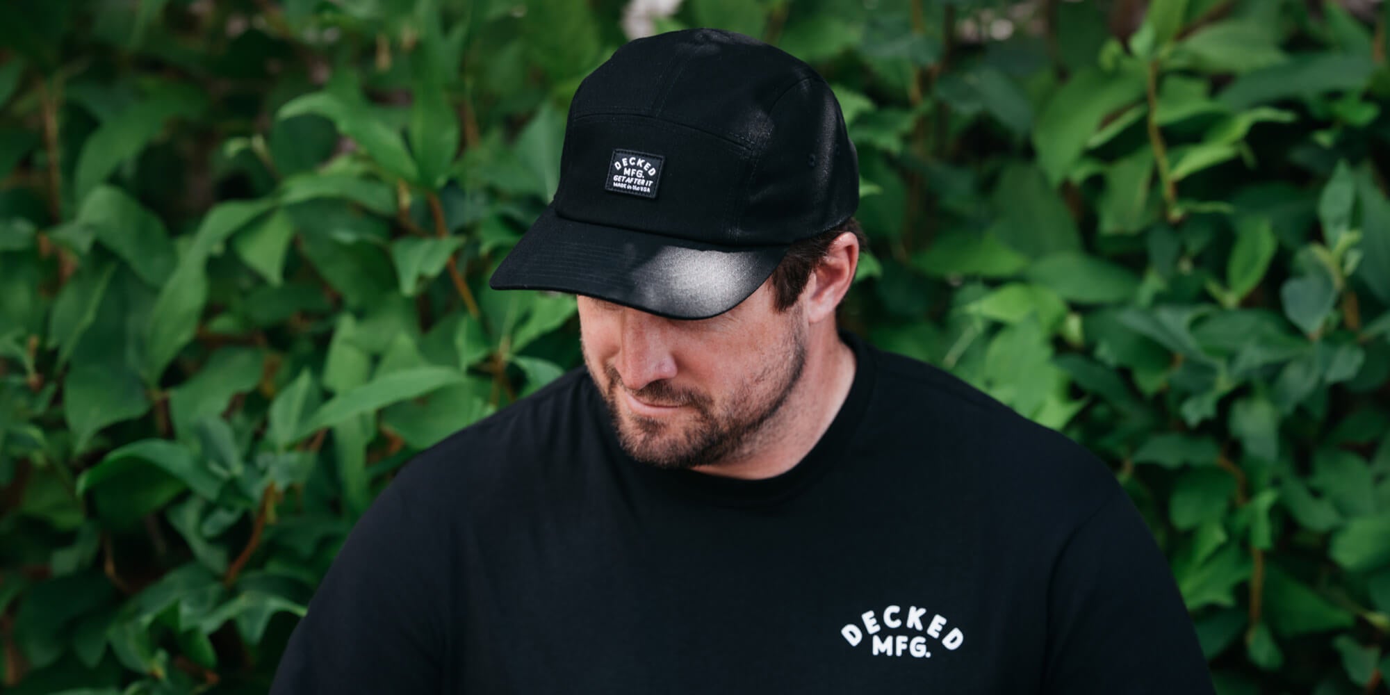 Man wearing a black cap and t-shirt with DECKED branding against a green leafy background
