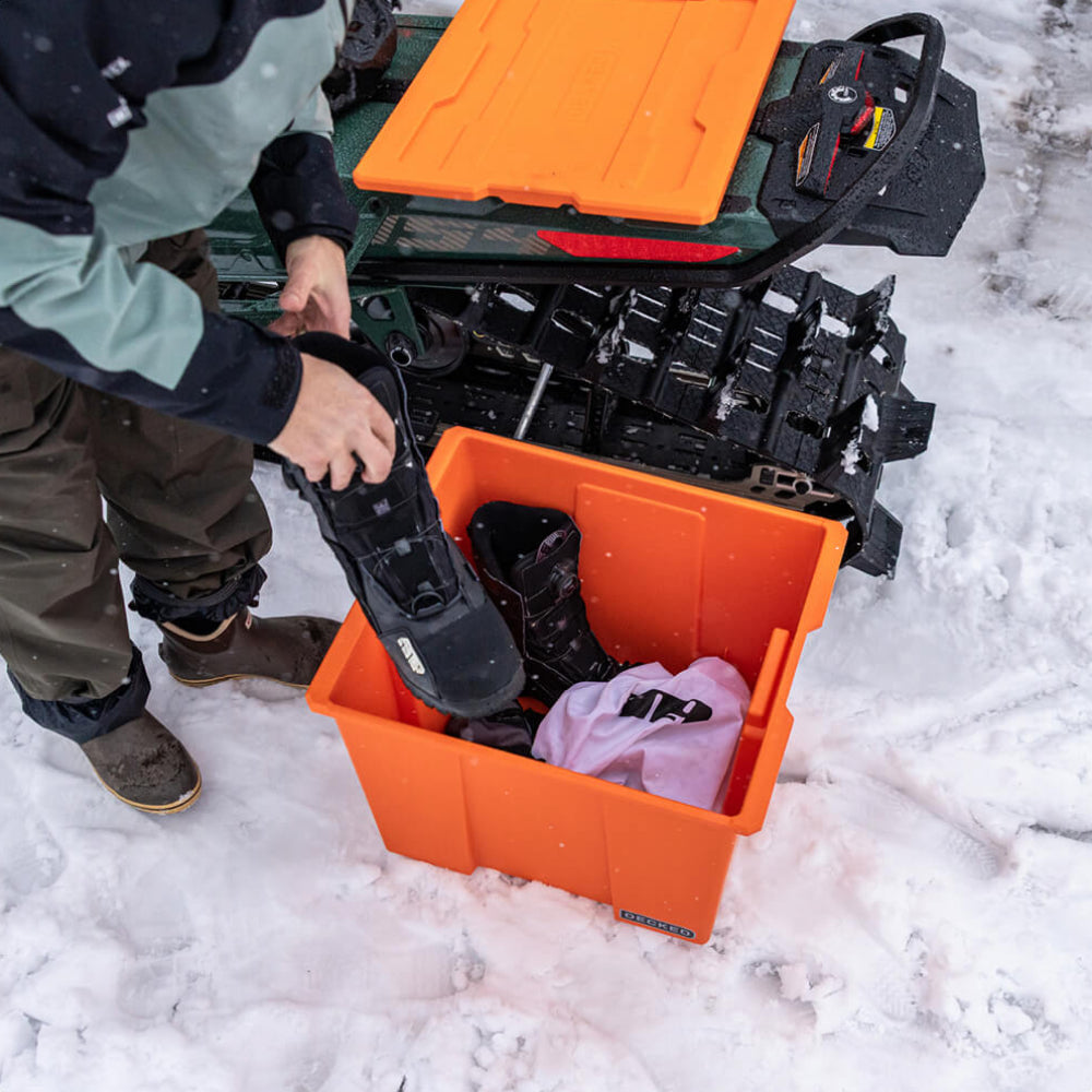 Person putting on a boot from an orange Payloader storage bin with snow equipment in the background
