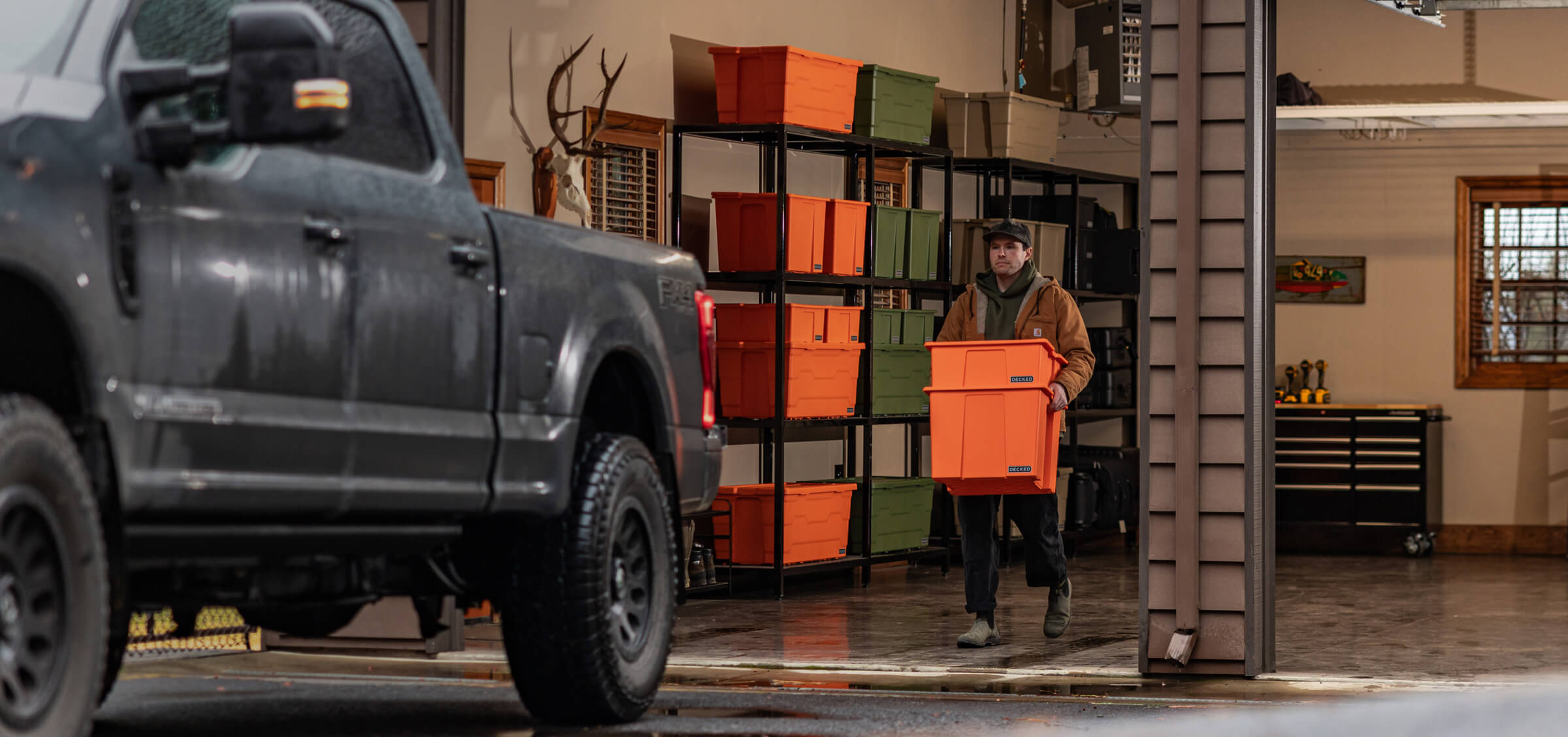 Person carrying two DECKED orange storage bins in a garage setting with a truck and storage shelves.