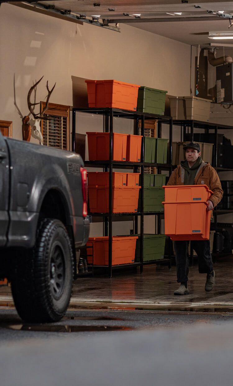 Person carrying two DECKED orange storage bins in a garage setting with a truck and storage shelves.