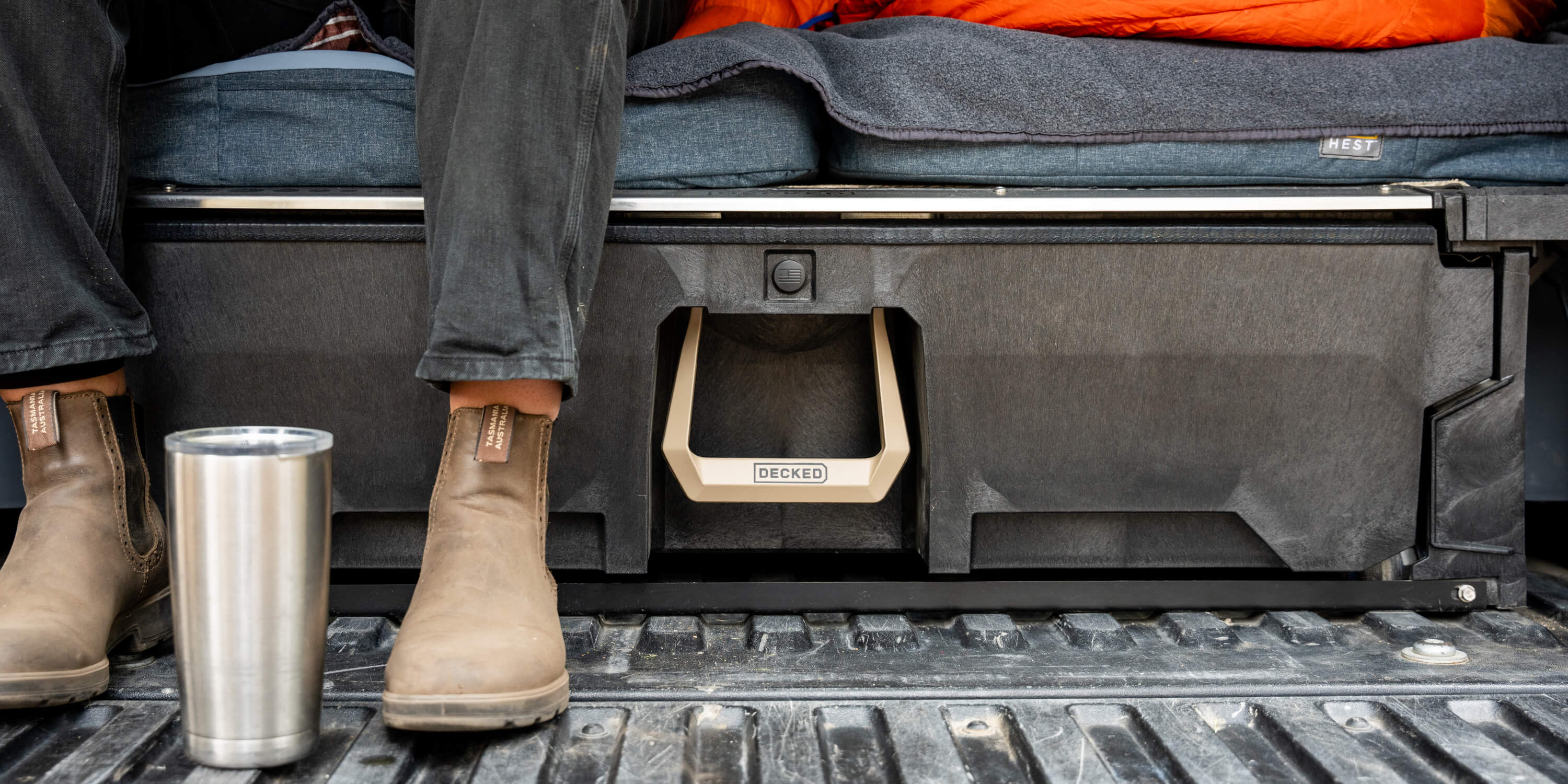 A woman sitting on her DECKED Drawer System, adorned with the exclusive Desert Tan Hop Up Kit. She's enjoying a cup of coffee and illustrating that she uses her DECKED Drawer System as a sleeping platform in the bed of her midsize truck.