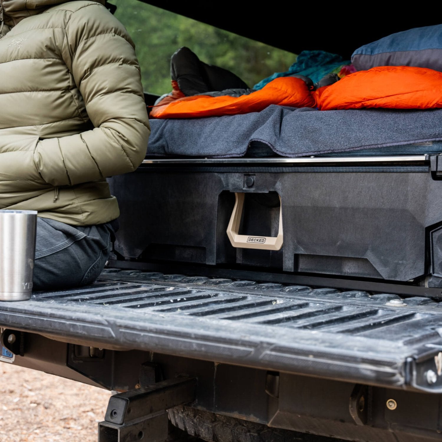 A woman sitting on her DECKED Drawer System, adorned with the exclusive Desert Tan Hop Up Kit. She's enjoying a cup of coffee and illustrating that she uses her DECKED Drawer System as a sleeping platform in the bed of her midsize truck.