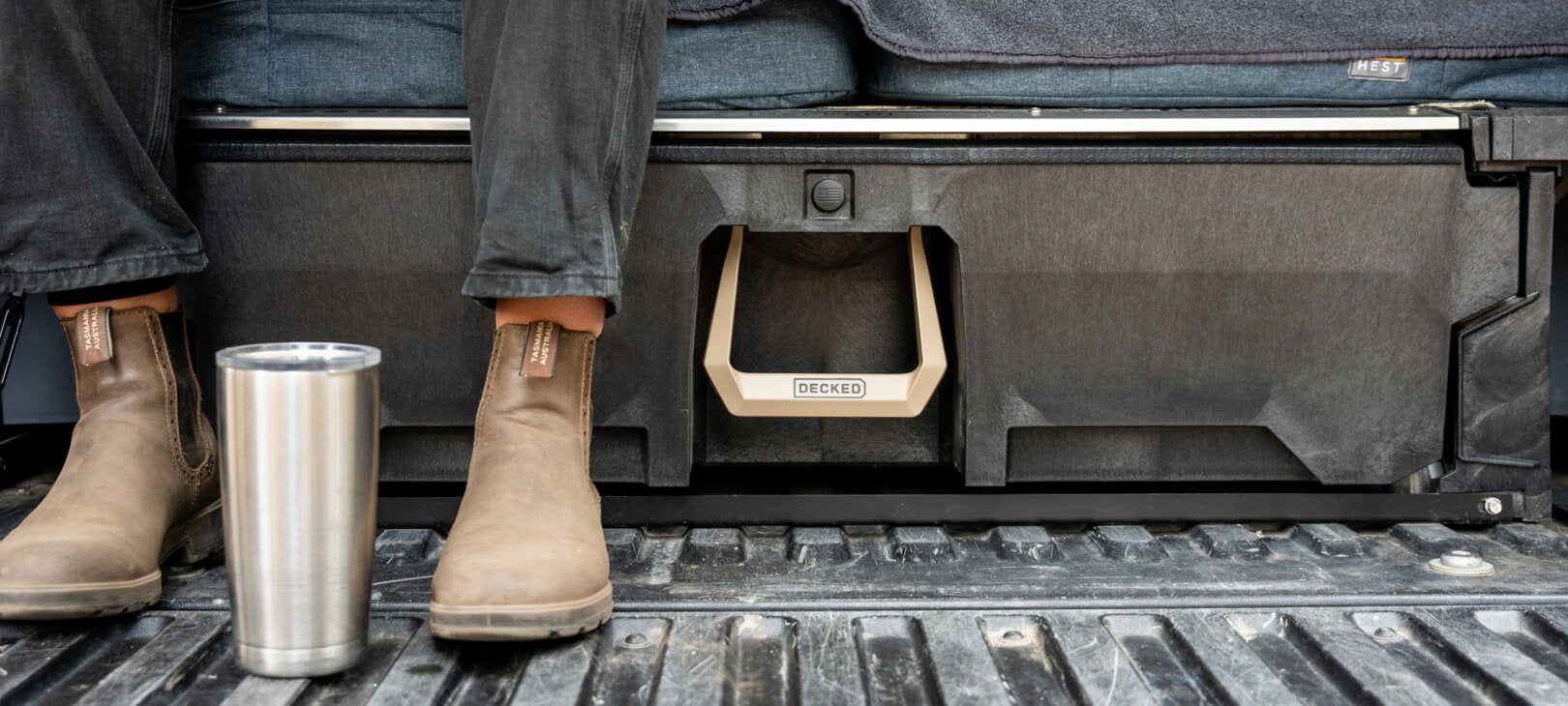 A woman sitting on her DECKED Drawer System, adorned with the exclusive Desert Tan Hop Up Kit. She's enjoying a cup of coffee and illustrating that she uses her DECKED Drawer System as a sleeping platform in the bed of her midsize truck.