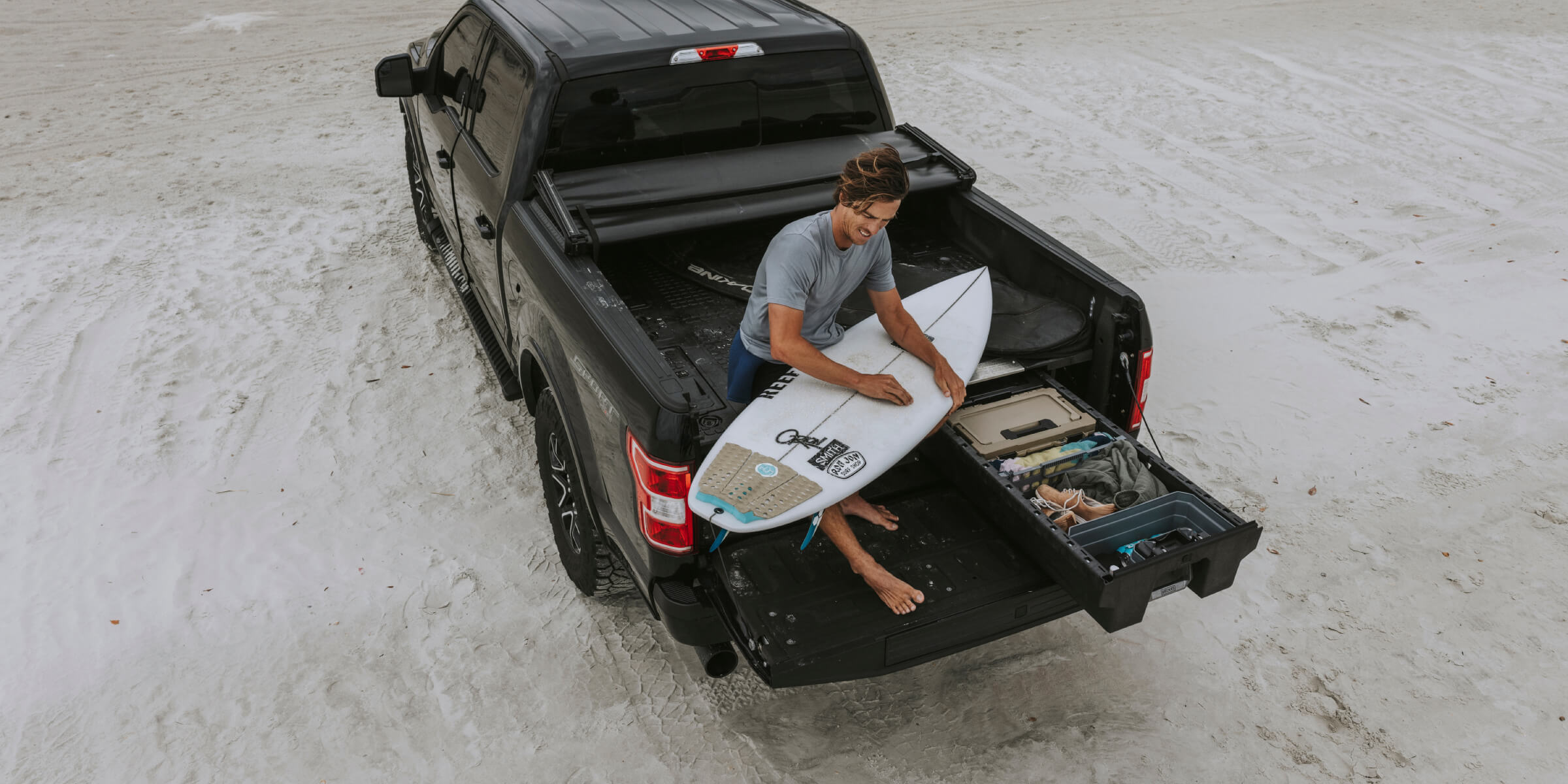A man waxing his surfboard on the beach, sitting on his DECKED Drawer System in the bed of his truck.