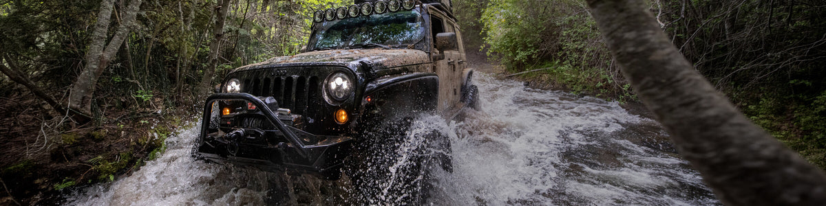 A mud-splattered black Jeep Wrangler with its headlights on drives through a rushing forest creek, water spraying off the front tire. The rig is built for overlanding, with a roof rack, a row of auxiliary lights above the windshield, and a steel front bumper with a winch. Dense green trees frame the narrow trail.