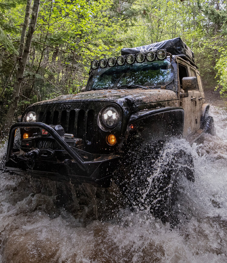A mud-splattered black Jeep Wrangler with its headlights on drives through a rushing forest creek, water spraying off the front tire. The rig is built for overlanding, with a roof rack, a row of auxiliary lights above the windshield, and a steel front bumper with a winch. Dense green trees frame the narrow trail.