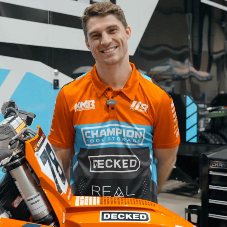 Kevin Moranz wearing an orange and black shirt with various brand logos, standing next to a motorcycle.