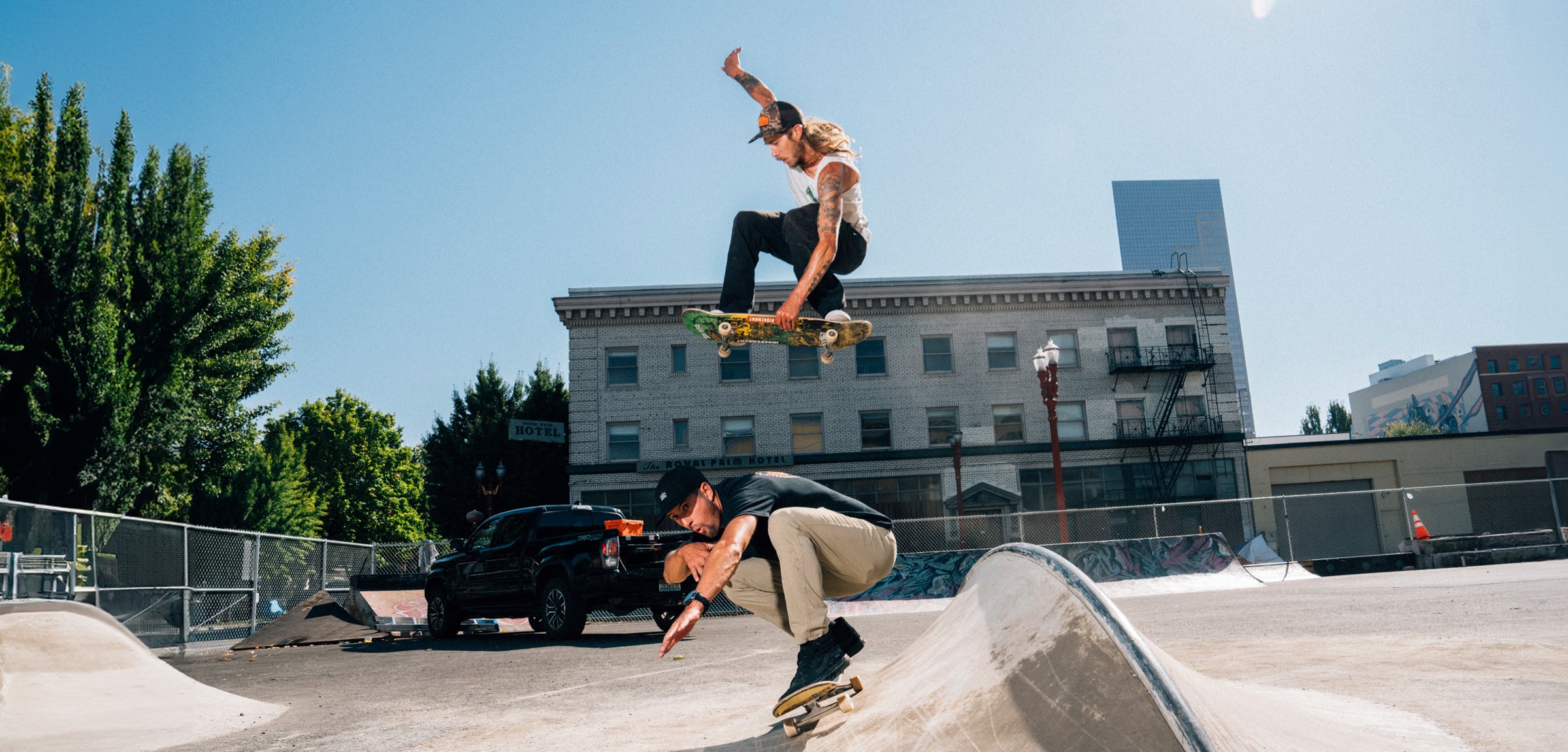 Two skateboarders performing tricks at a skate park with buildings and trees in the background.