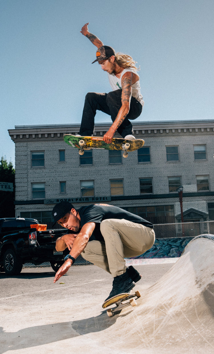 Two skateboarders performing tricks at a skate park with buildings and trees in the background.