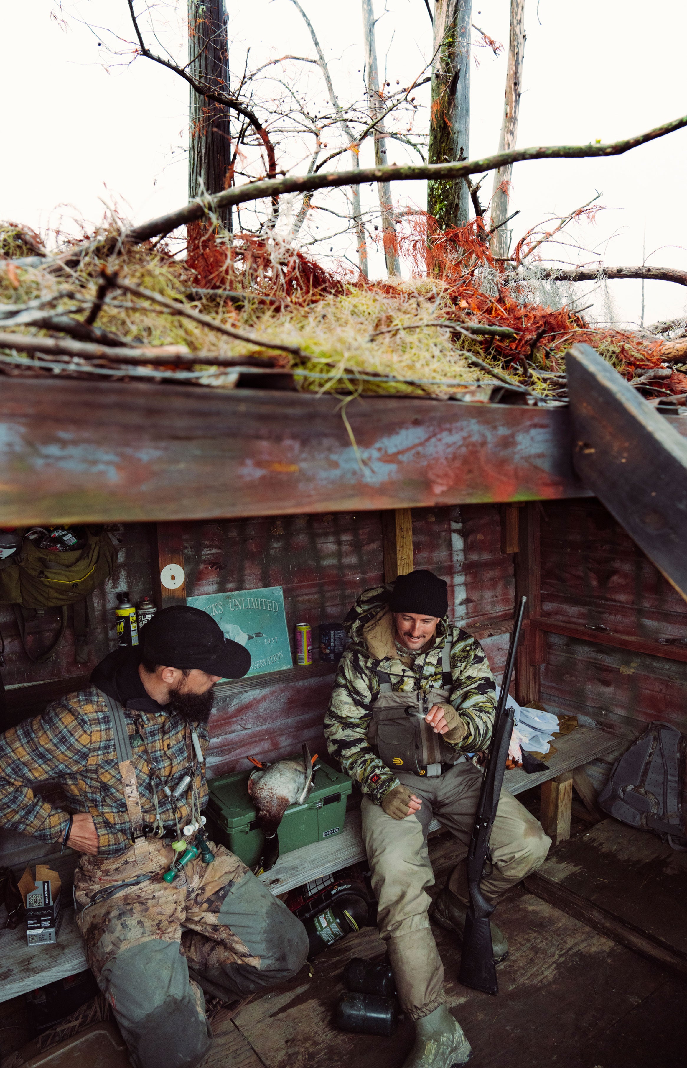 Two hunters in camouflage gear sitting inside a wooden blind in a forest setting.