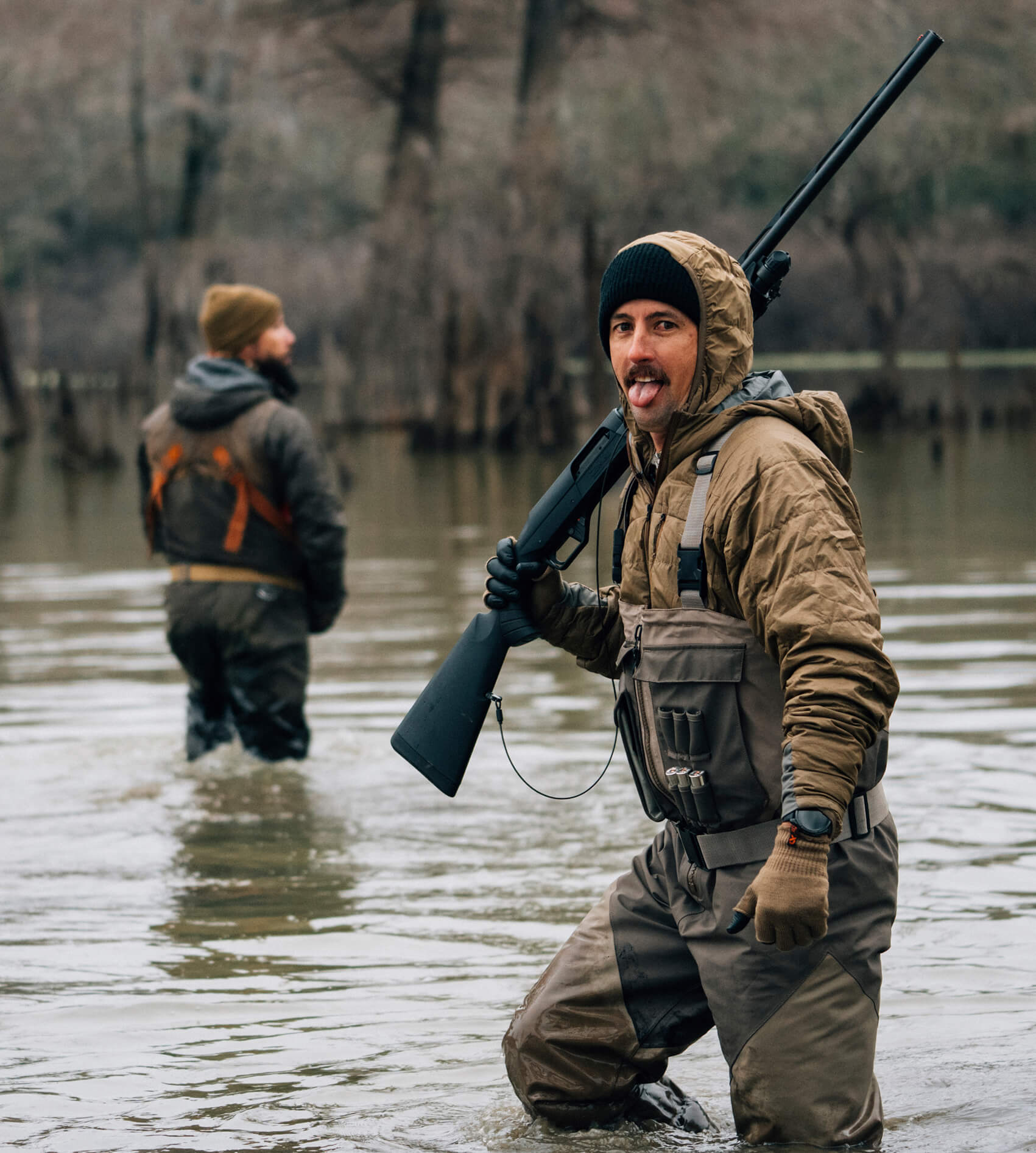Man in waders holding a rifle in a flooded area with another person in the background.