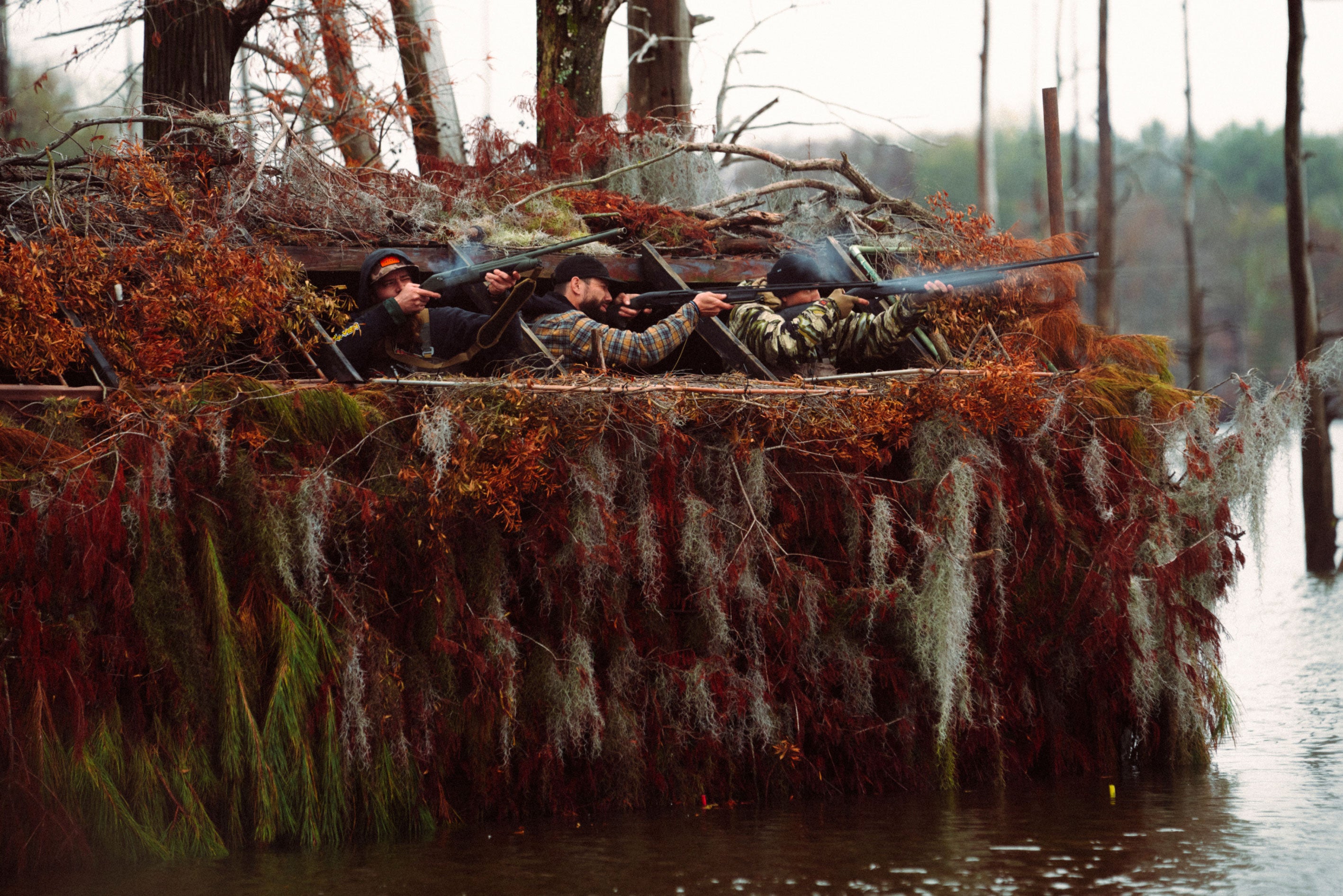 Three hunters in camouflage gear on a duck blind in a marshy area.
