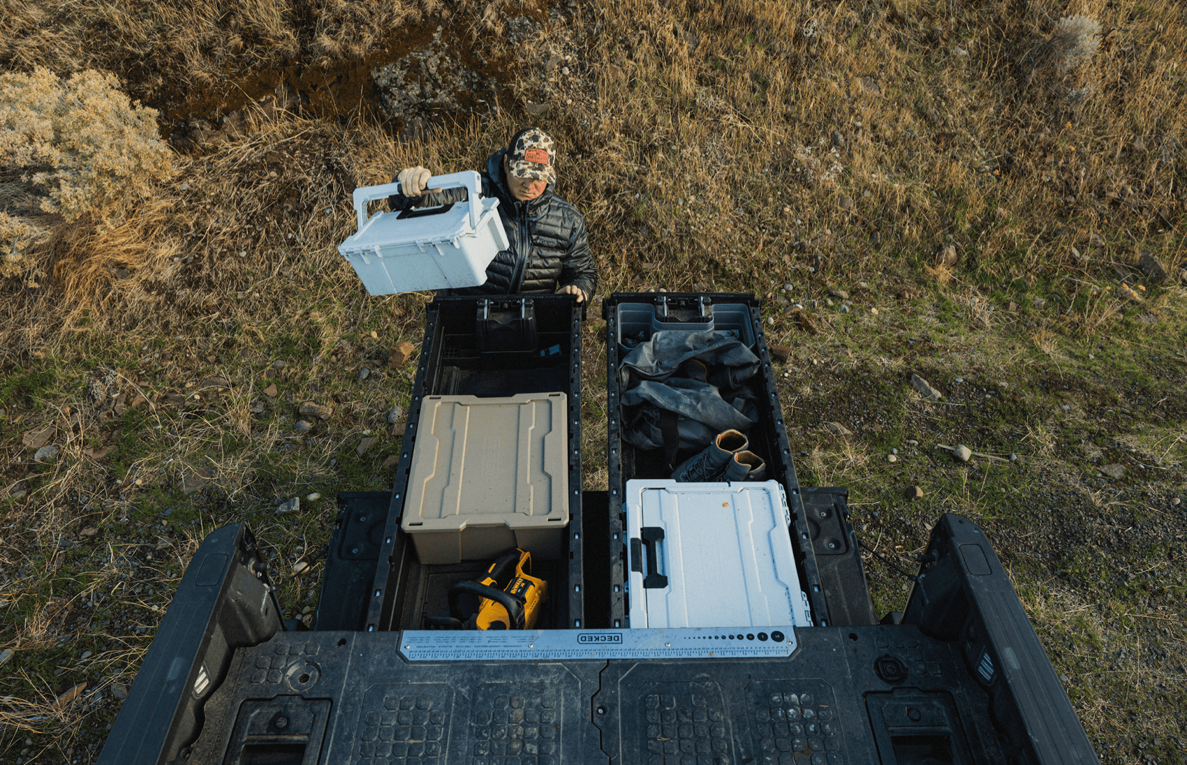 Person in camouflage holding a container in a vehicle with tools and equipment on a grassy field.