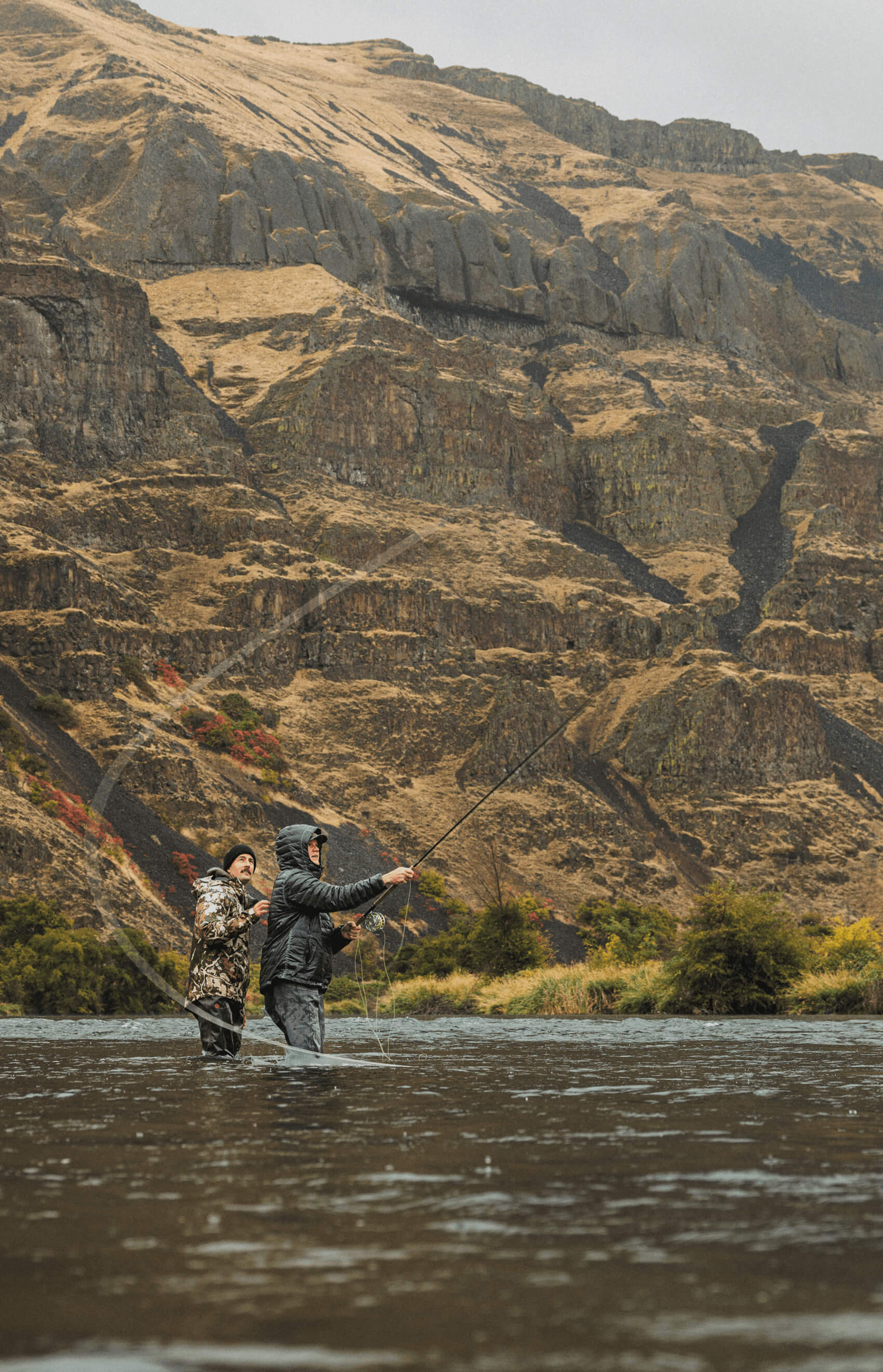 Two people fly fishing in a river with a mountainous landscape in the background