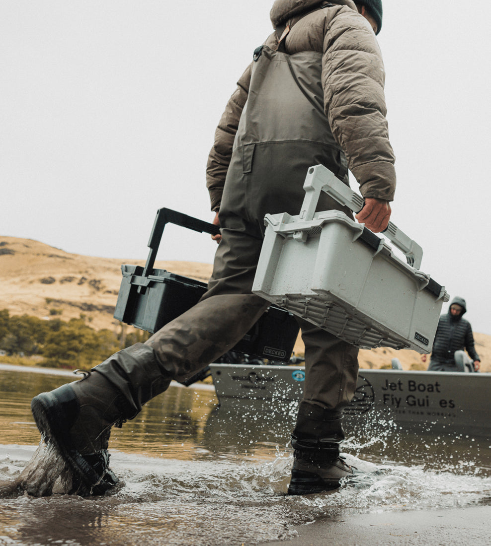 Person carrying a large gray case near a body of water with mountains in the background.
