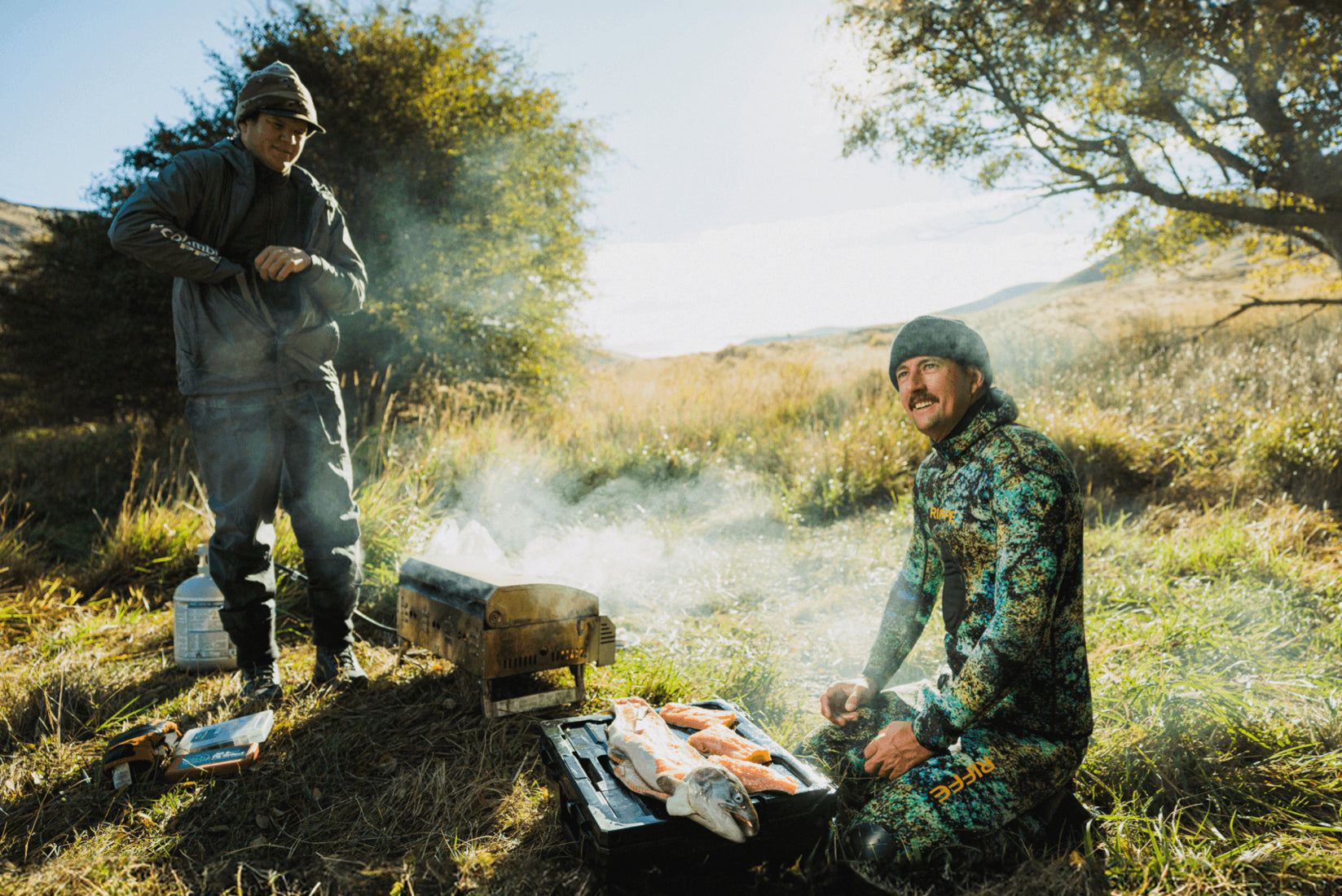 Two men cooking outdoors in a natural setting with smoke rising from a grill.