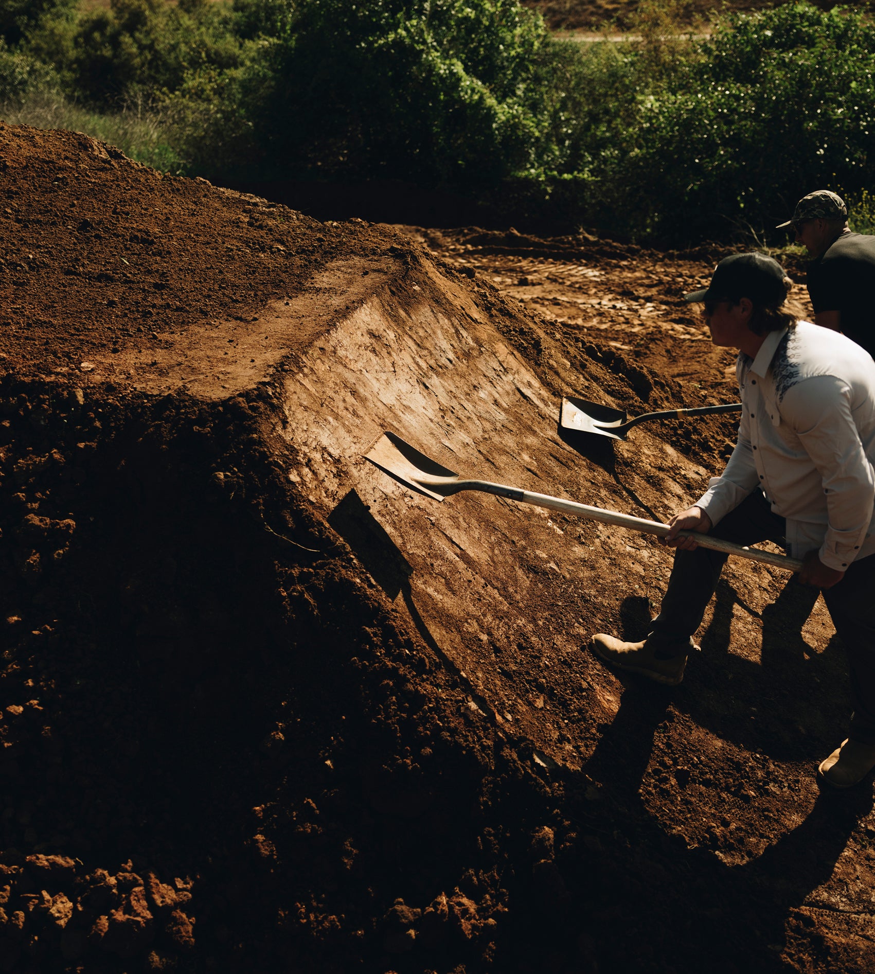 Kyle Strait and Danny Bolton working with shovels in a dirt area with greenery in the background