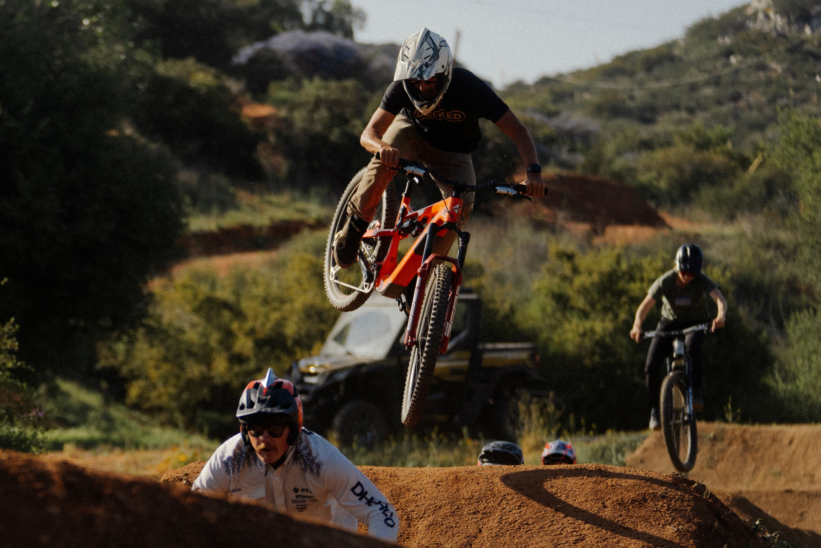 Danny Bolton in mid-air during a jump on a dirt track with other bikers and vehicles in the background.