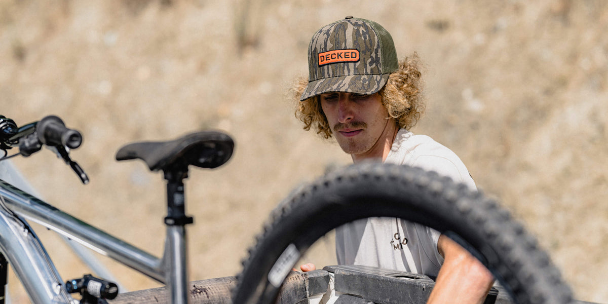 Young man with curly hair wearing a Mossy Oak camo DECKED trucker hat, working on a mountain bike outdoors.