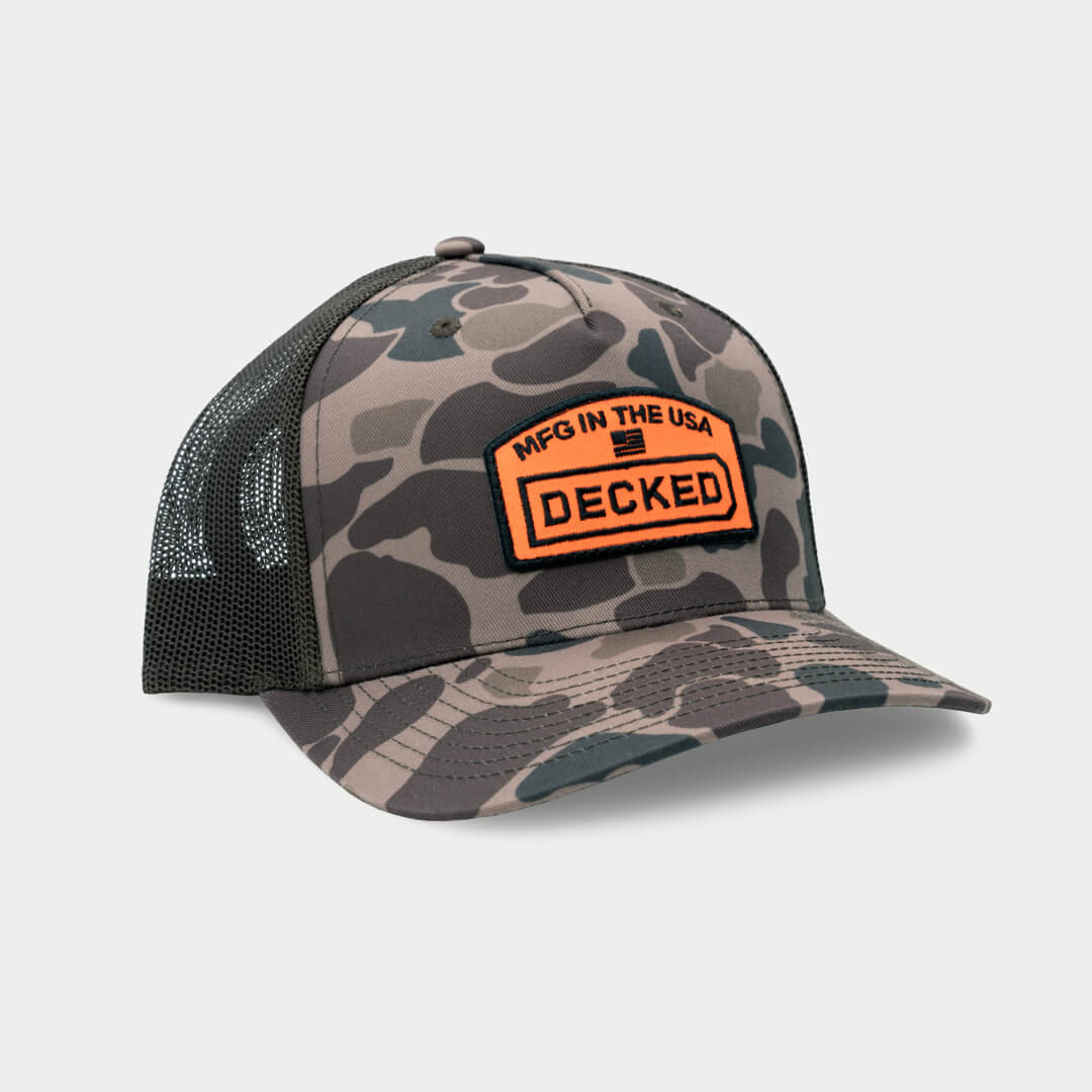 A green and brown camo hat with a mesh back with an orange badge on it that has an embroidered "MFG in the USA", American Flag, and DECKED logo.