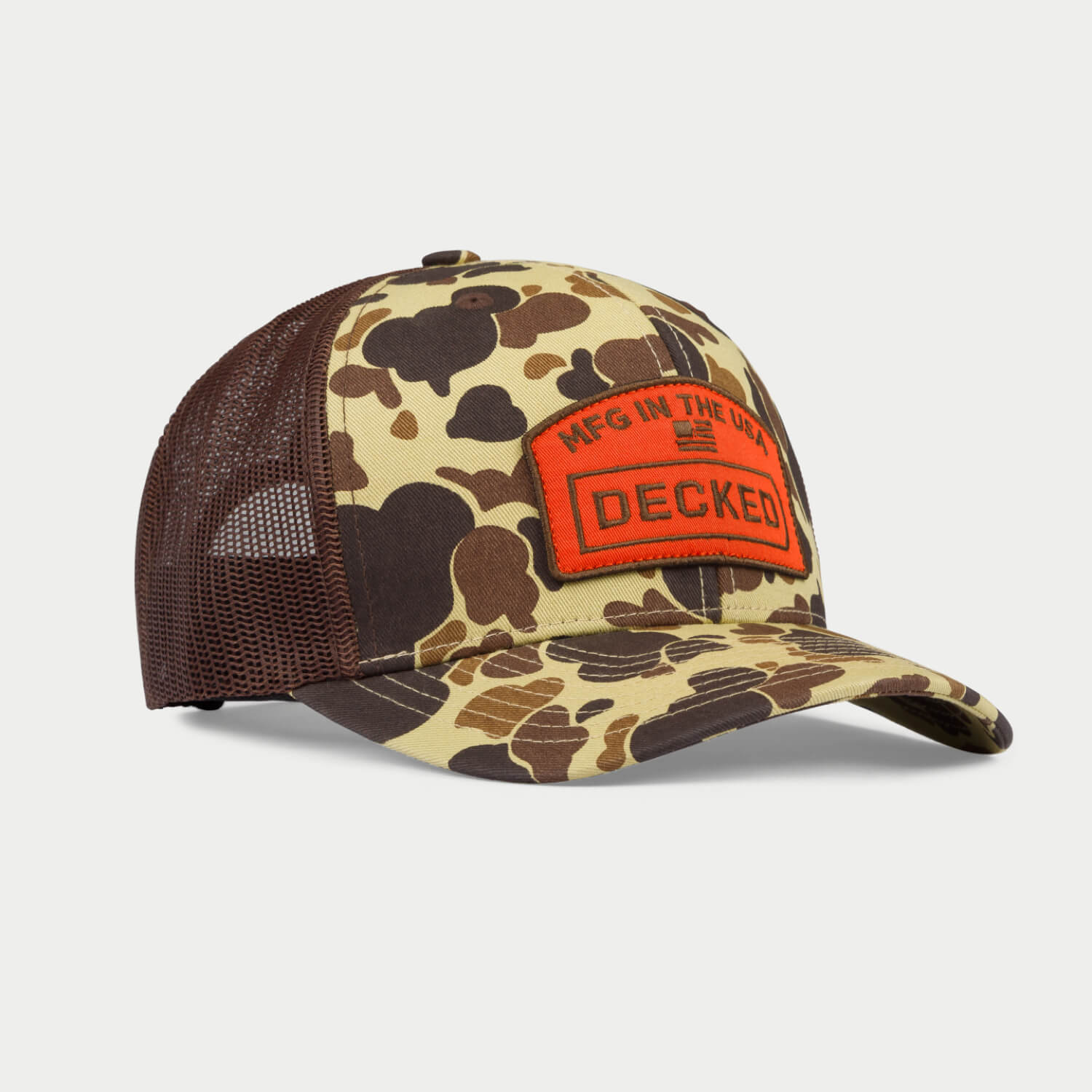  [H0112-HATR-CMO]  An angled green and brown camo hat with a mesh back with an orange badge on it that has an embroidered "MFG in the USA", American Flag, and DECKED logo.