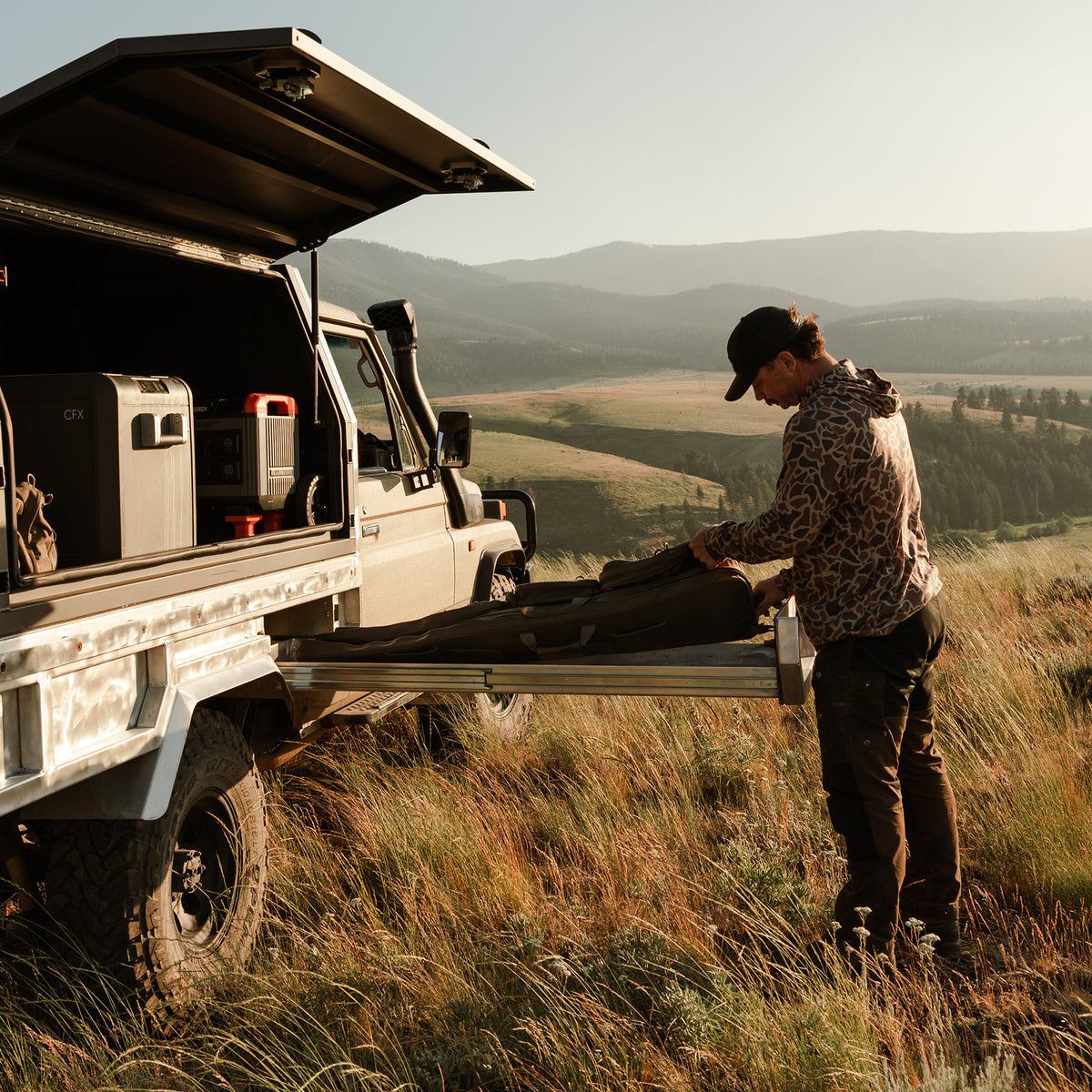 Man unloading outdoor gear from a truck-mounted storage system using a slide-out cargo tray in a grassy mountain landscape.