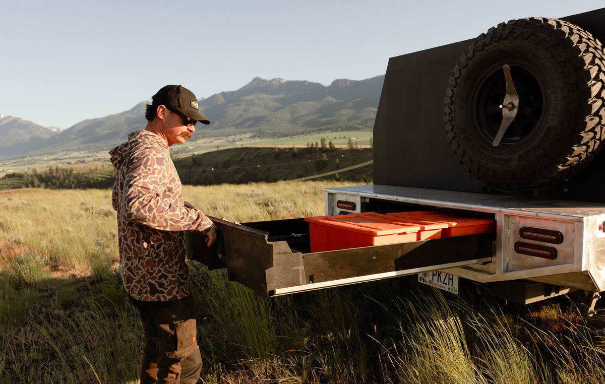 Man opening a DECKED truck drawer system filled with orange DECKED sixer bins on a Toyota Land Cruiser, parked in a grassy mountain landscape.