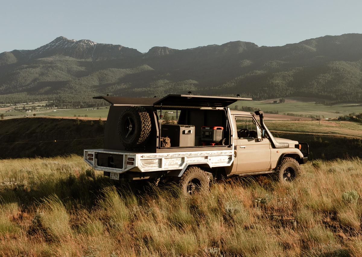 1999 Toyota Land Cruiser equipped with a DECKED truck storage system, parked in a grassy field with the rear storage compartments open, surrounded by mountain scenery.