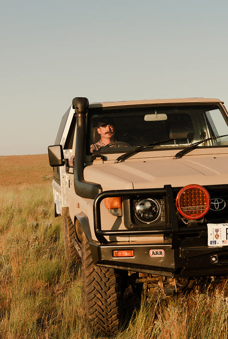A beige Toyota Land Cruiser off-road vehicle with a black snorkel, front bull bar, and round auxiliary light, driven by a man in a grassy field under a clear sky.