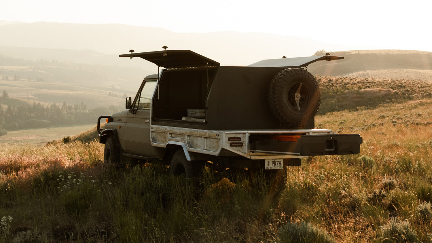 DECKED truck storage system mounted in the bed of a pickup truck parked in a grassy mountain landscape, with both side panels open and a spare tire mounted on the rear.